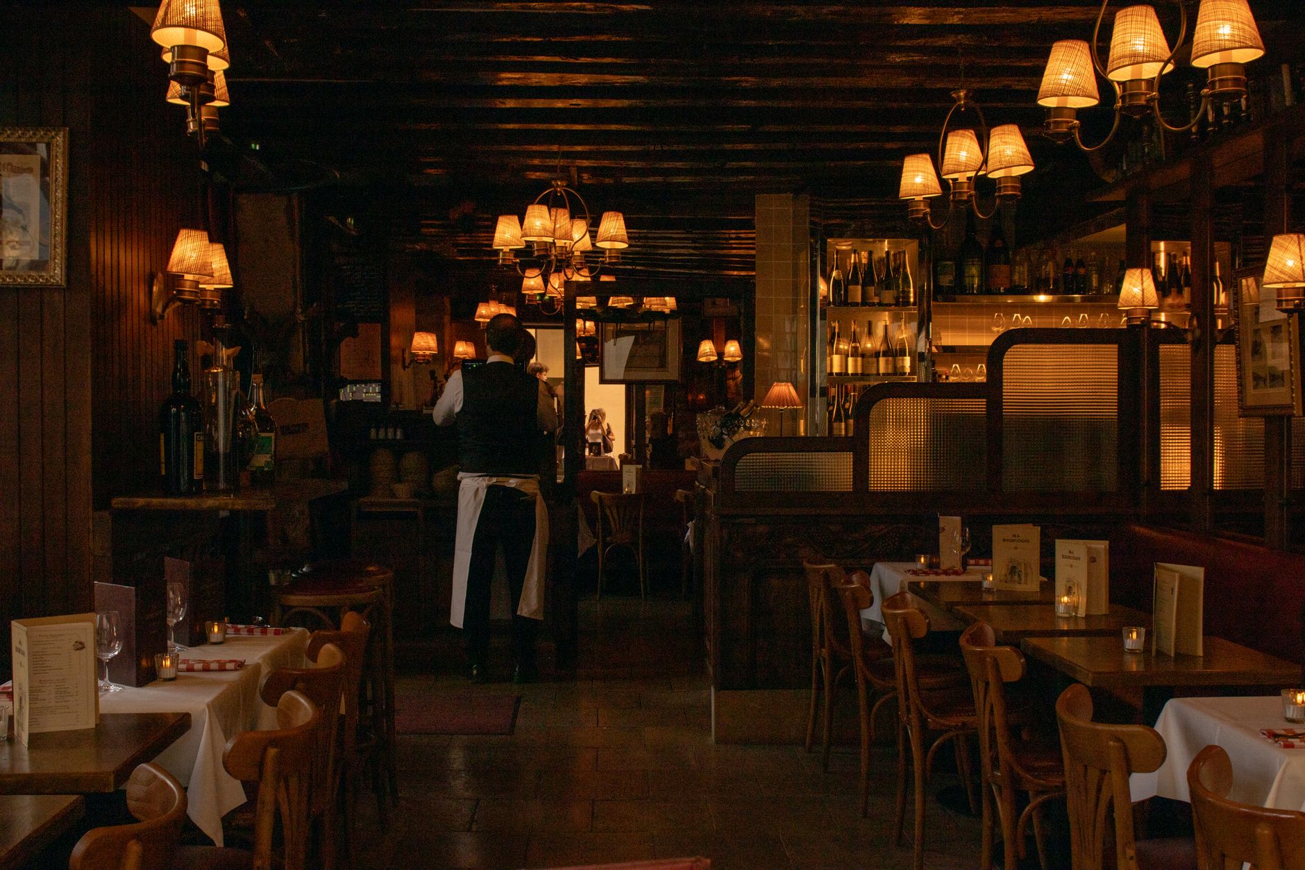 Nima & Nonna's dining room in the evening, with closely set wooden tables, low pendant lights and a long banquette against a plastered wall